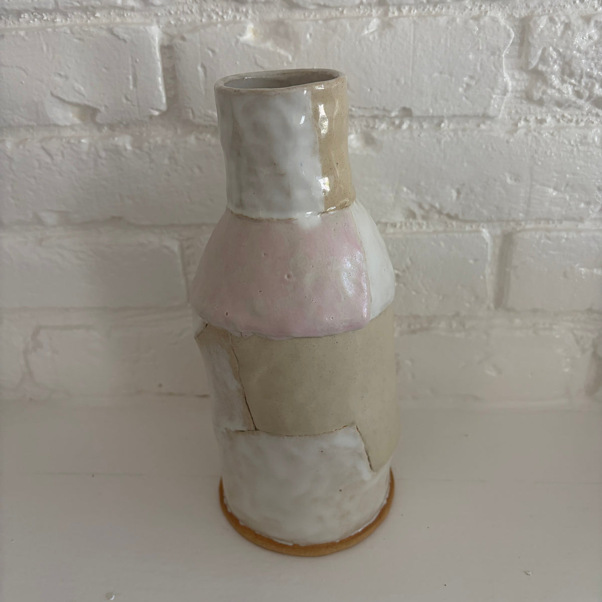 A handmade ceramic vase with a textured, patchwork-style glaze in soft pastel shades of pink, beige, and off-white, standing on a white surface against a white brick wall.