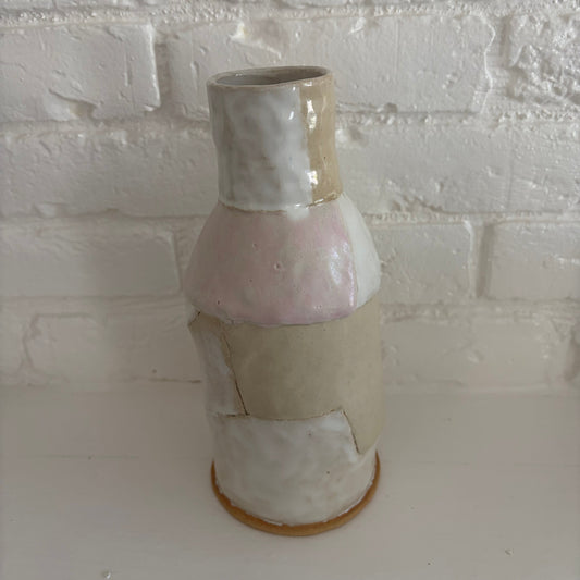 A handmade ceramic vase with a textured, patchwork-style glaze in soft pastel shades of pink, beige, and off-white, standing on a white surface against a white brick wall.