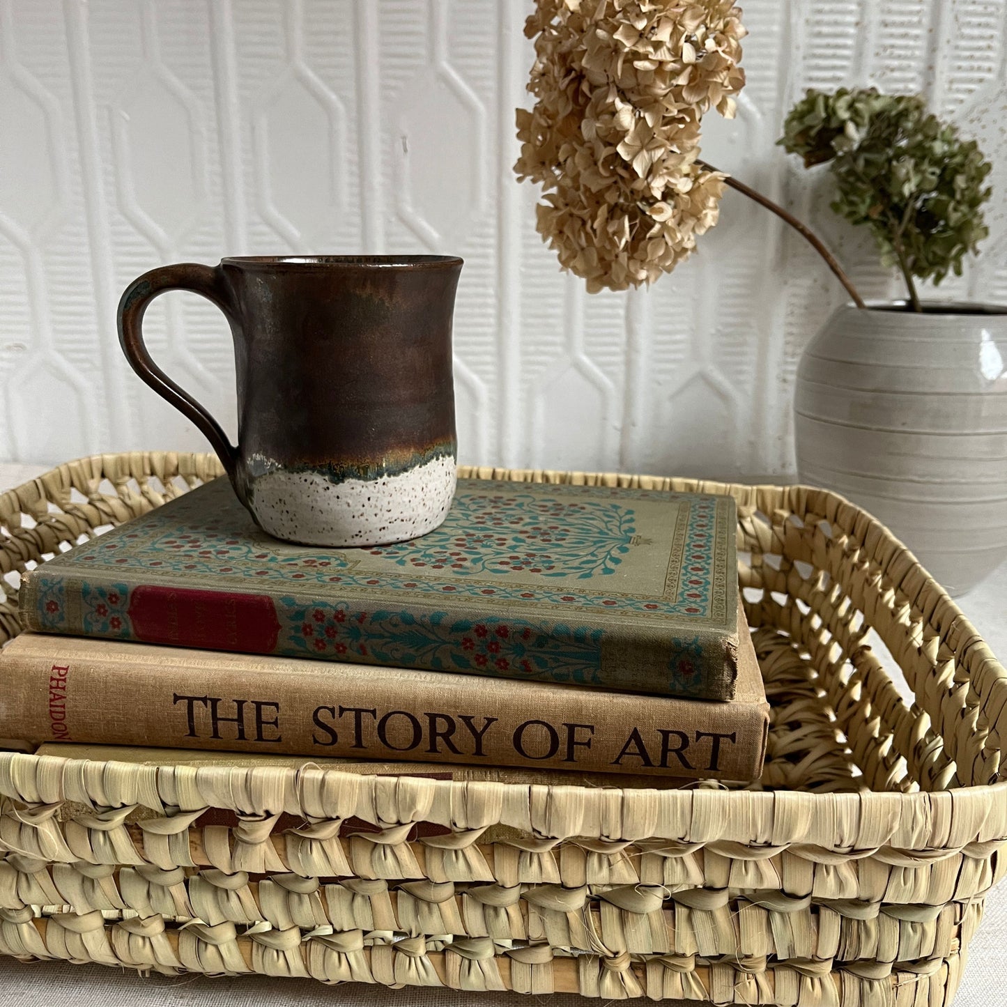 Ceramic mug on top of books in a woven basket with decorative plants in the background