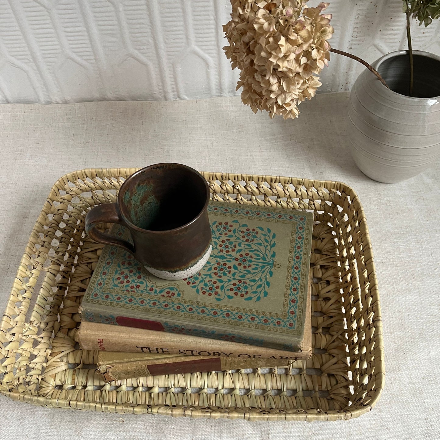 Woven tray with books and a mug on a light surface, with a plant in the background.