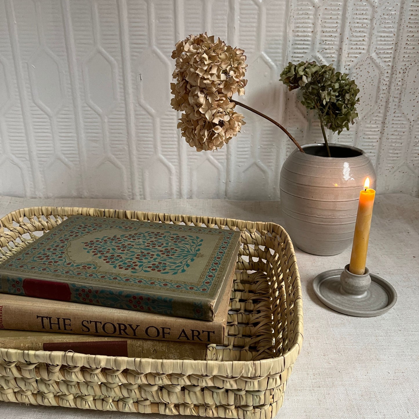 Woven basket with books, vase with flowers, and candle on a textured surface.