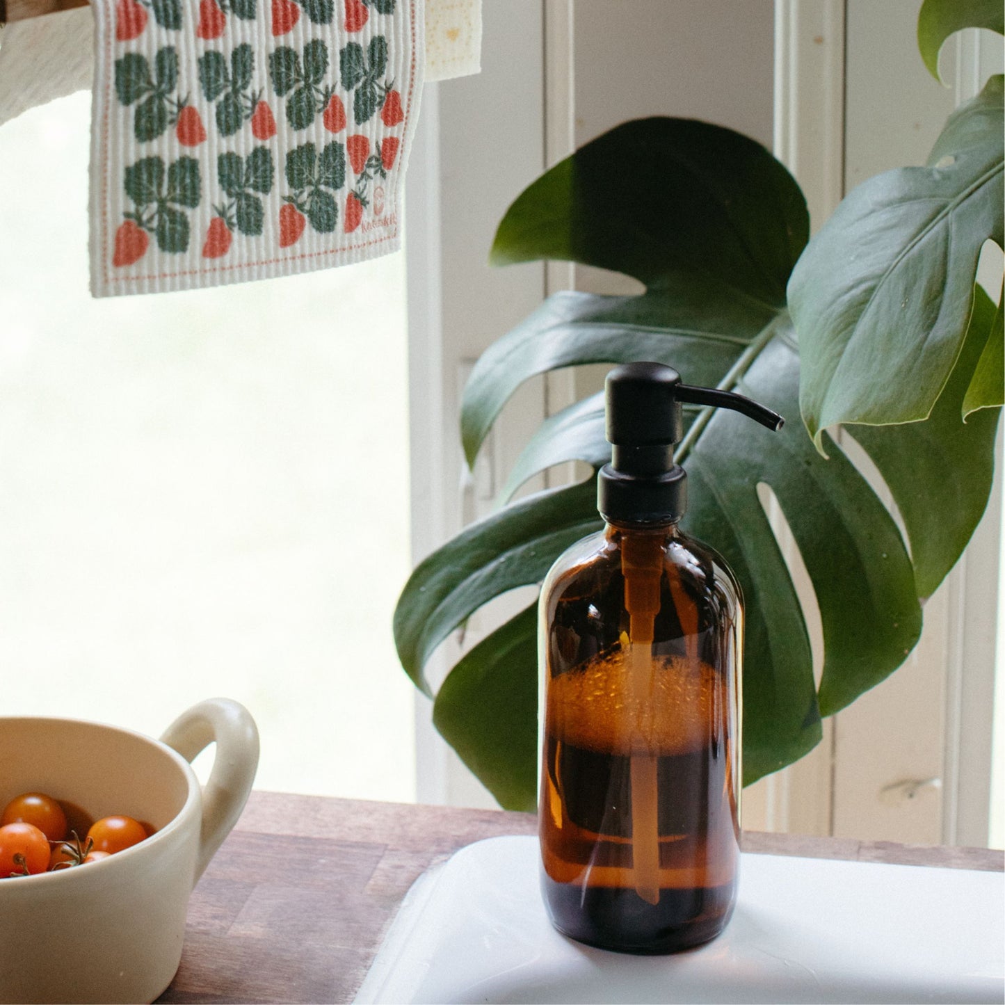 Reusable Amber Glass Soap Dispenser  with a bowl filled with tomatoes, a dishcloth placed above and a plant in the back