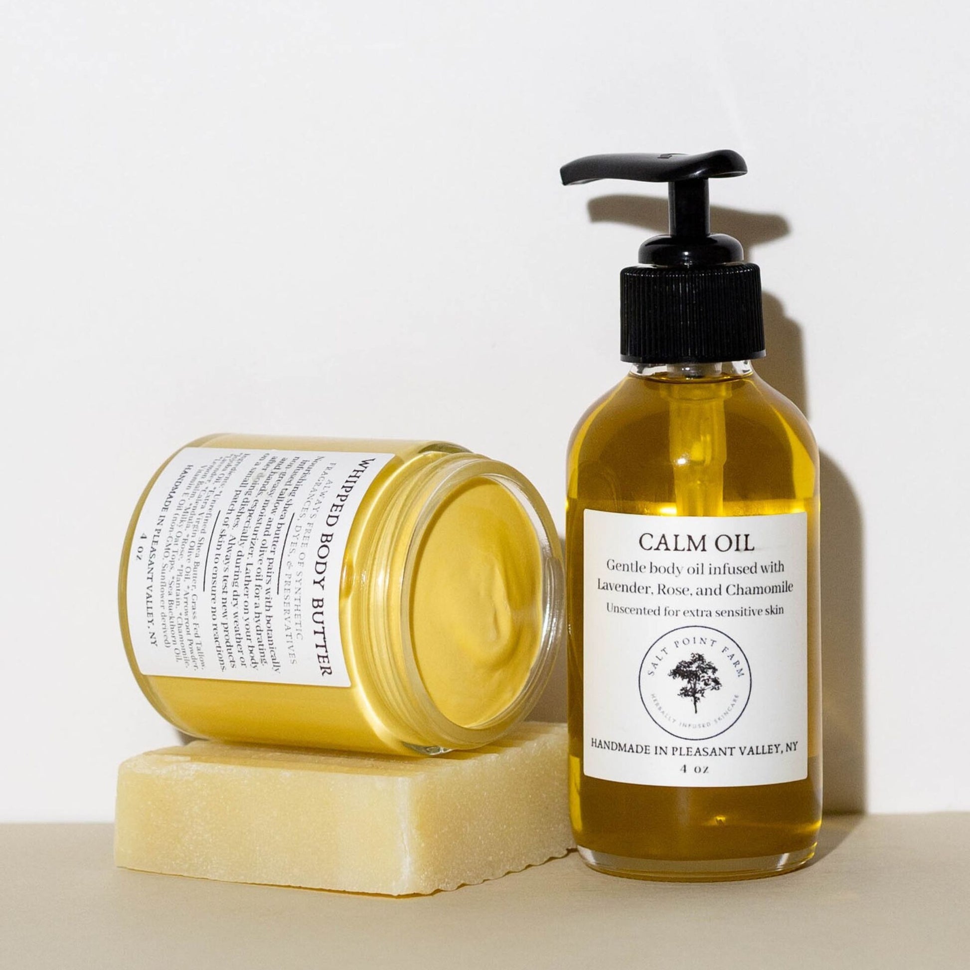 Bottle of calm Body Oil with Dispenser next to a Whipped Body Butter placed on a piece of soap.