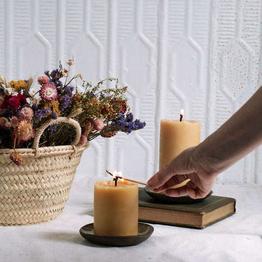 Person lighting a candle next to a basket of flowers and a stack of books on a textured white wall background.