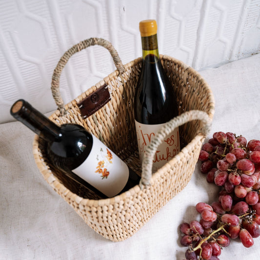 Woven basket with two wine bottles and a bunch of grapes on a light surface.