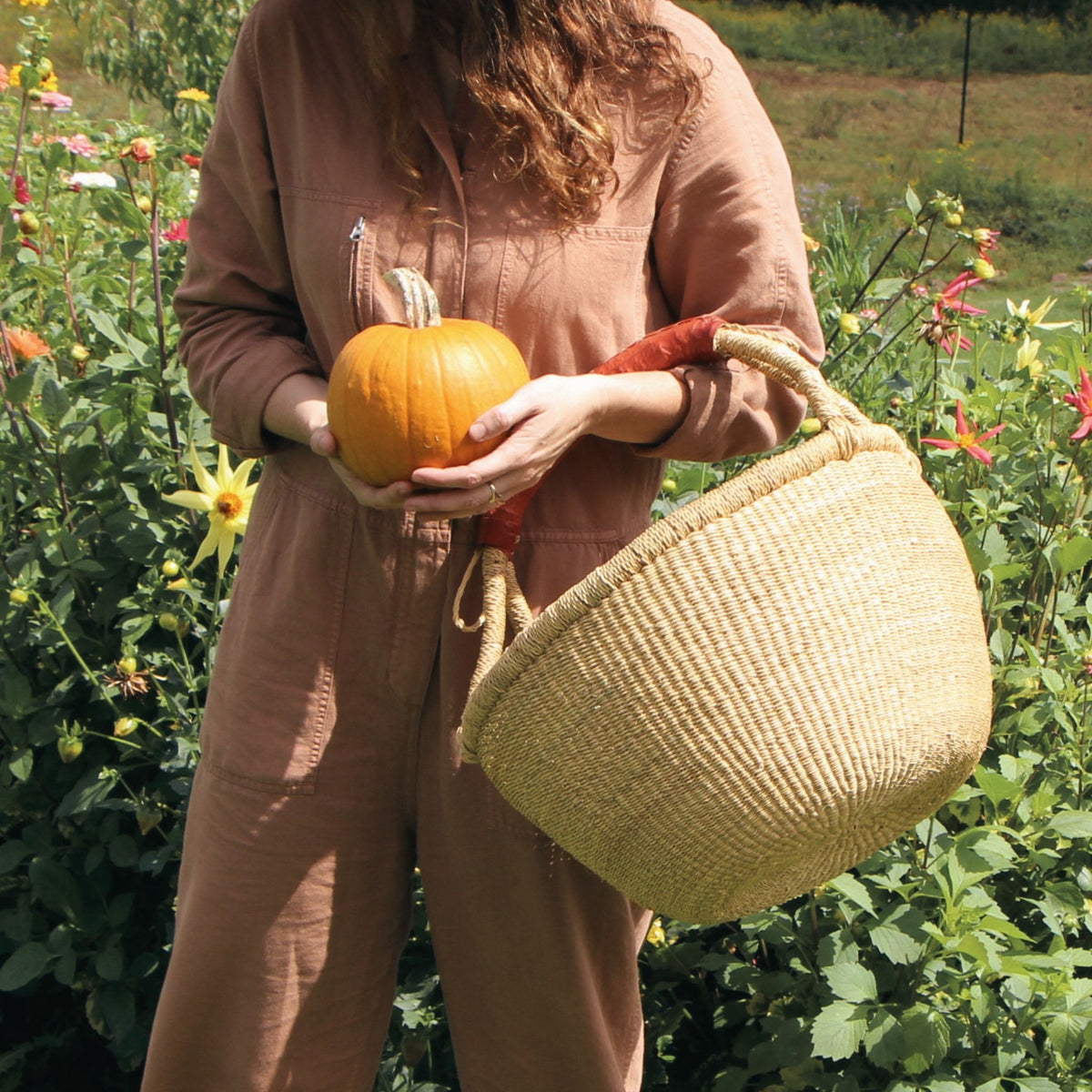 A person outdoors in a flower garden holds a small orange pumpkin and carries a large woven straw basket with a red handle. The person is wearing a dusty rose-brown jumpsuit.