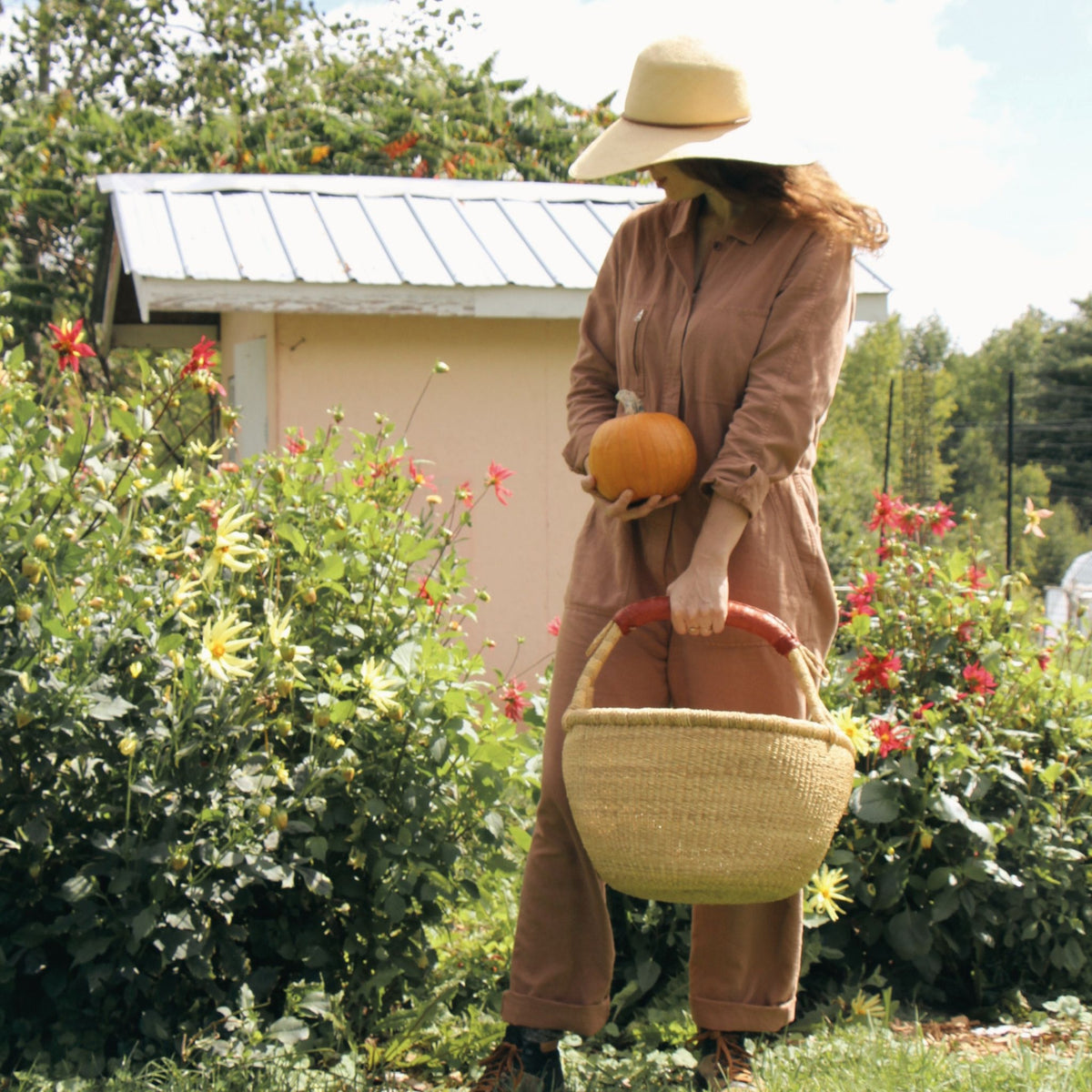 A woman in a brown linen jumpsuit and straw hat stands in a garden holding a small orange pumpkin and a woven straw basket with a dark red handle. A small shed and flowering bushes are in the background.