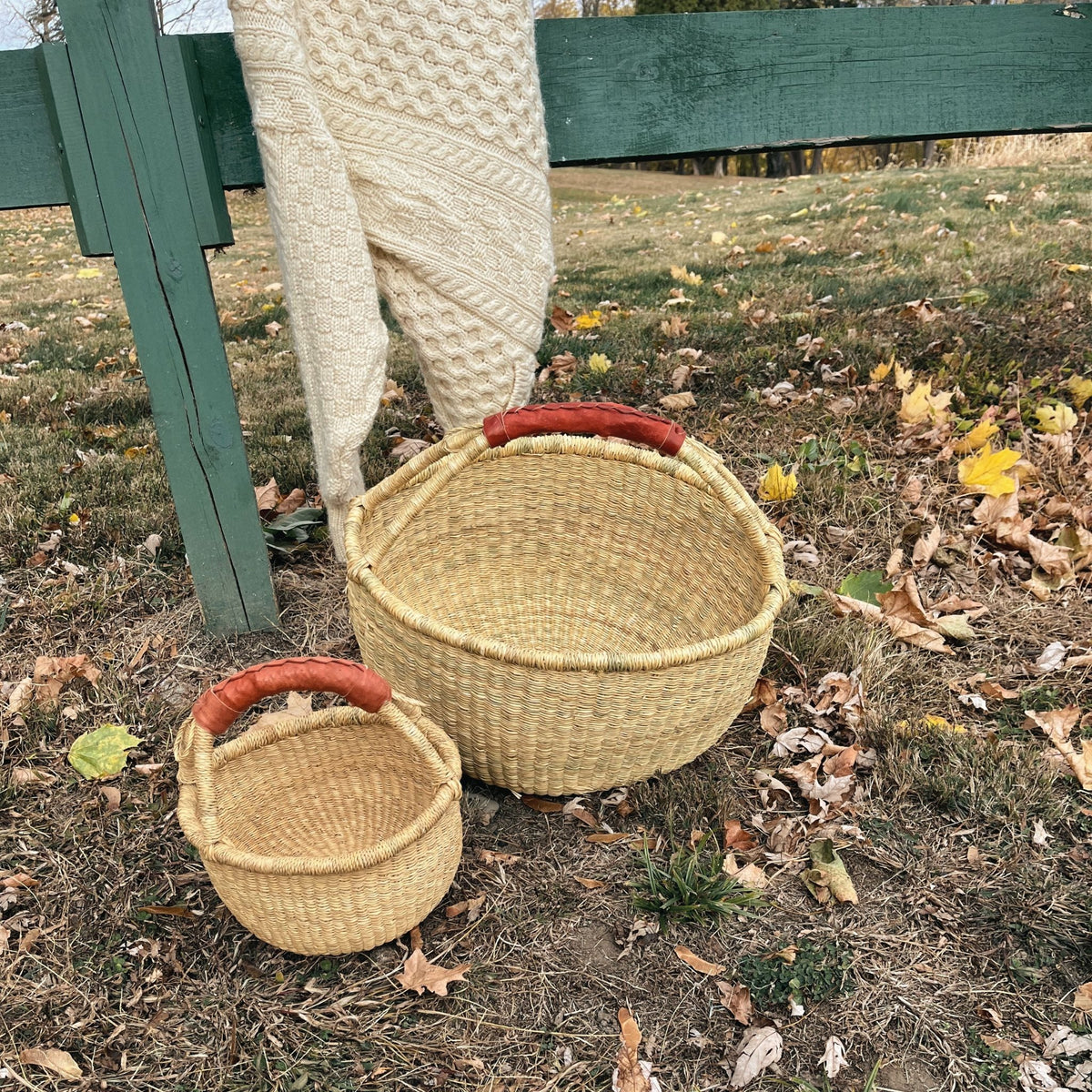 Two woven natural-fiber baskets with reddish-brown handles placed outdoors on grass among fallen leaves, with a green fence and a cream knit blanket in the background.
