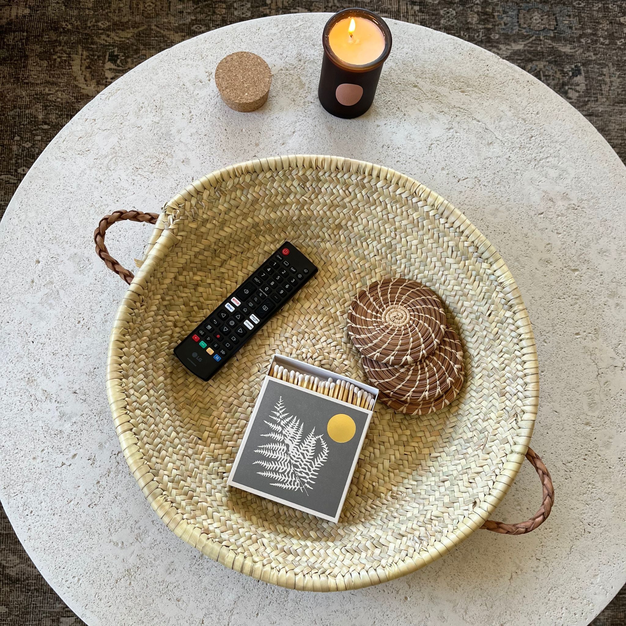 Woven tray with leather handles on a white circular table. On the table is a lit candle and inside the basket is a remote, box of matches, and coasters. 