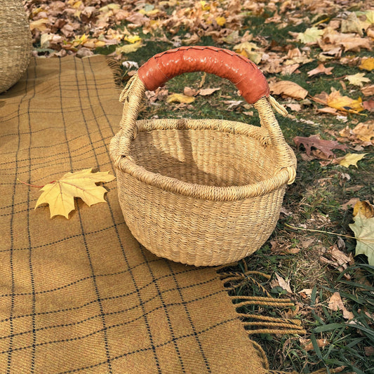 A woven straw basket with a curved orange-brown leather handle sits on a tan plaid blanket outdoors on grass covered with fallen autumn leaves.