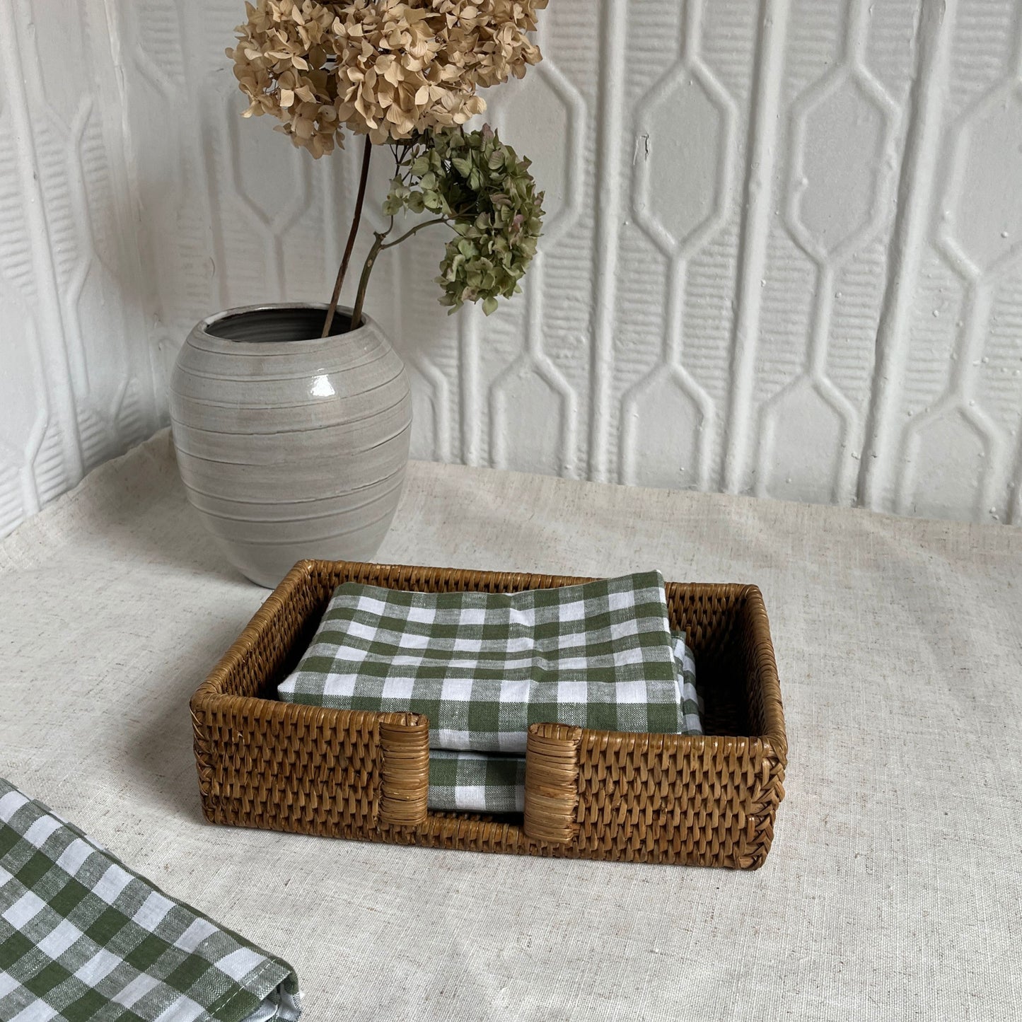 Woven basket with green checkered napkins on a textured surface with a decorative vase in the background.
