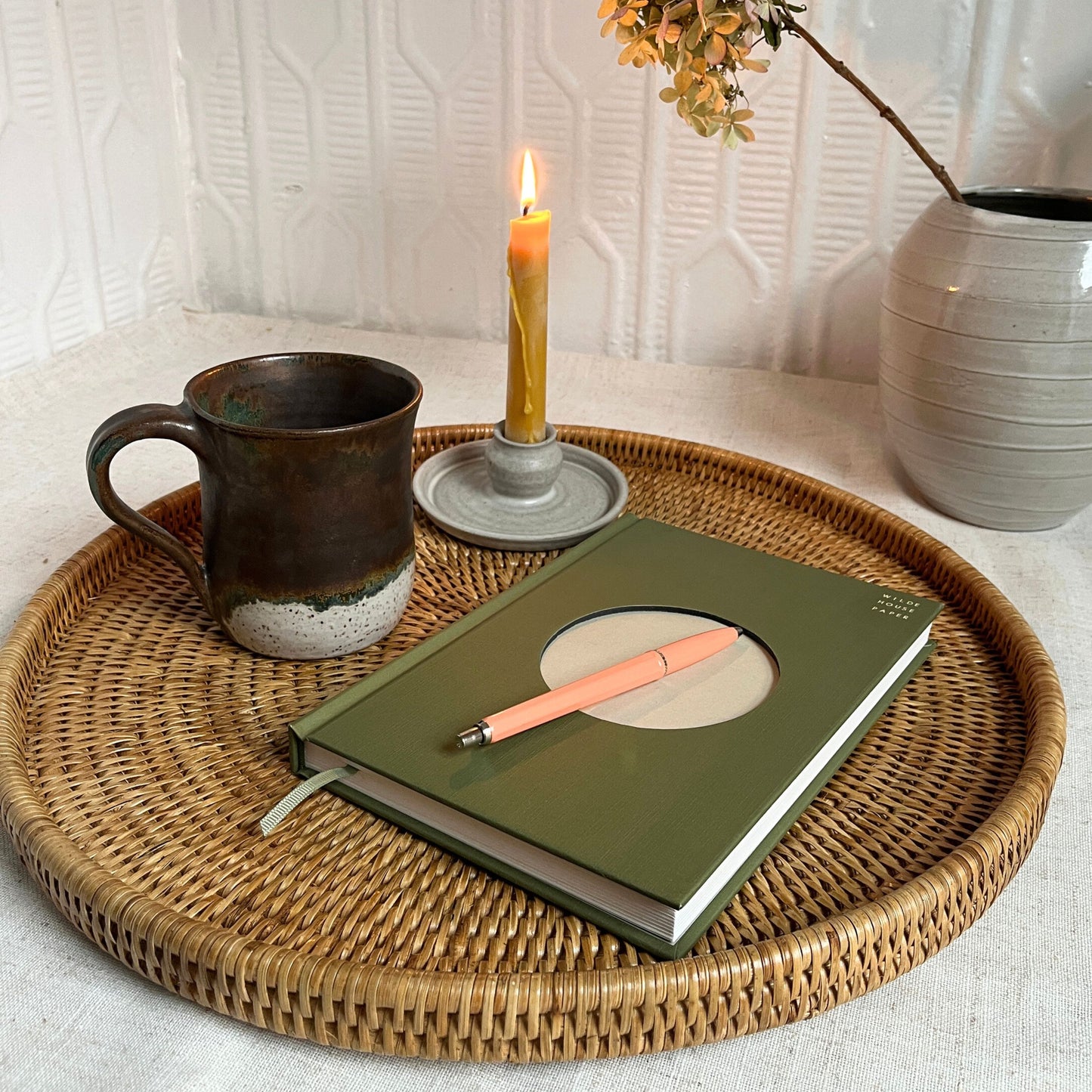 Green notebook with a pen on a woven tray with a mug, candle, and vase in the background.