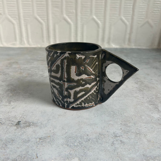 Black, green, white, and red patterned mug with a triangle handle. 