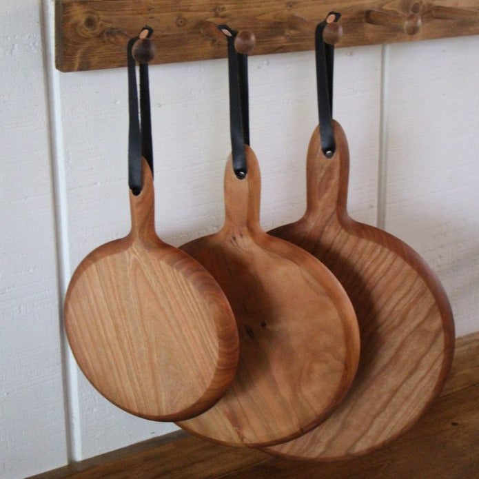X-Large, large, and medium circular cutting boards in cherry with handle and leather strap shown hanging from peg rail