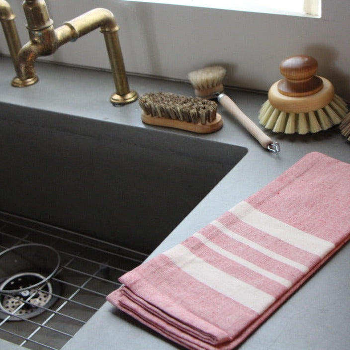 100% handwoven soft cotton kitchen towel in red tomato stripe with hanging loop, resting next to a sink and some cleaning brushes