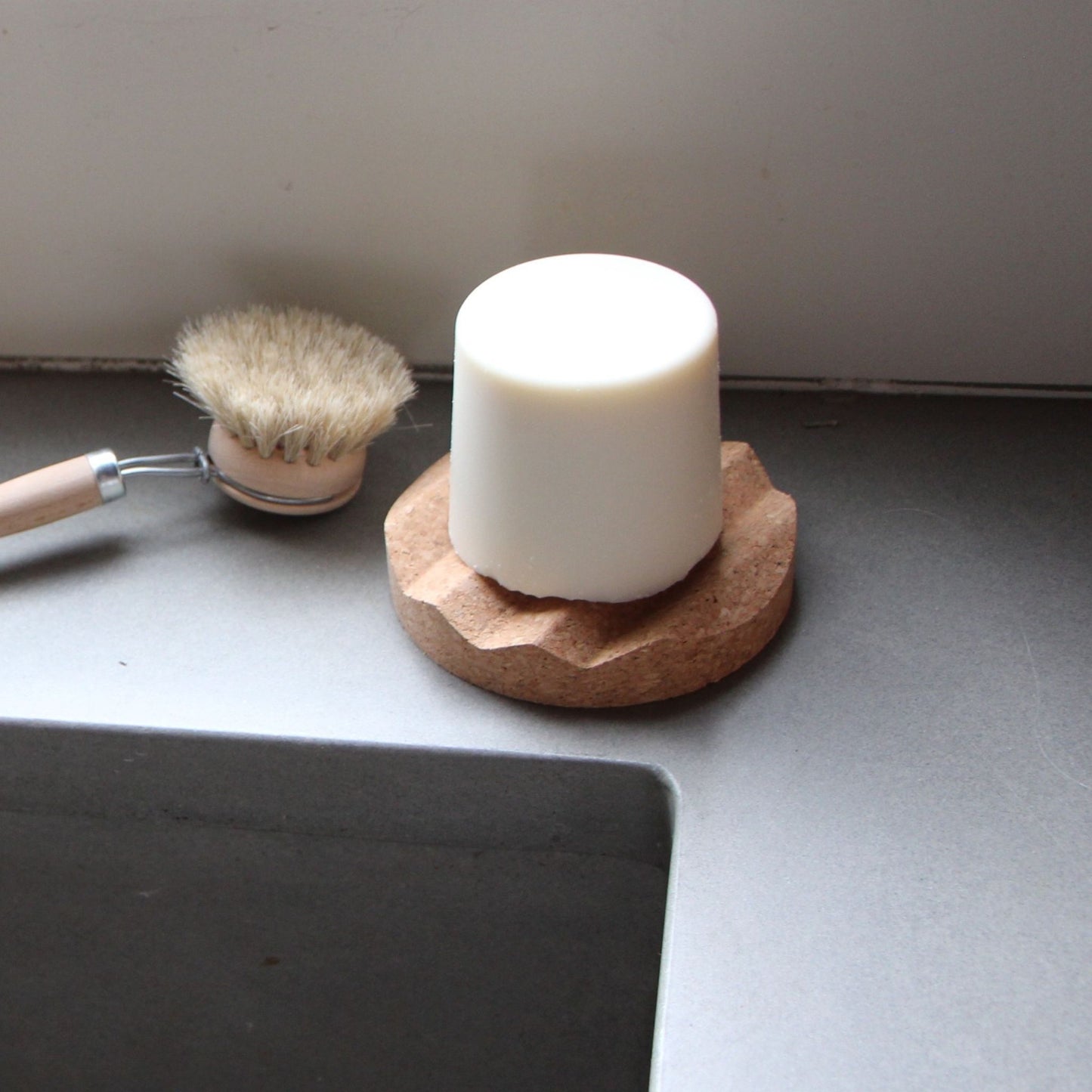 solid dish soap bar on a cork soap dish with a dish brush next to a sink