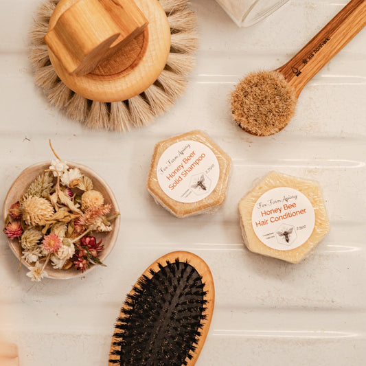 Two Honey Bee Conditioner, a little bowl of dried flowers, a hair brush, and two cleaning brushes from different sizes. All natural wood