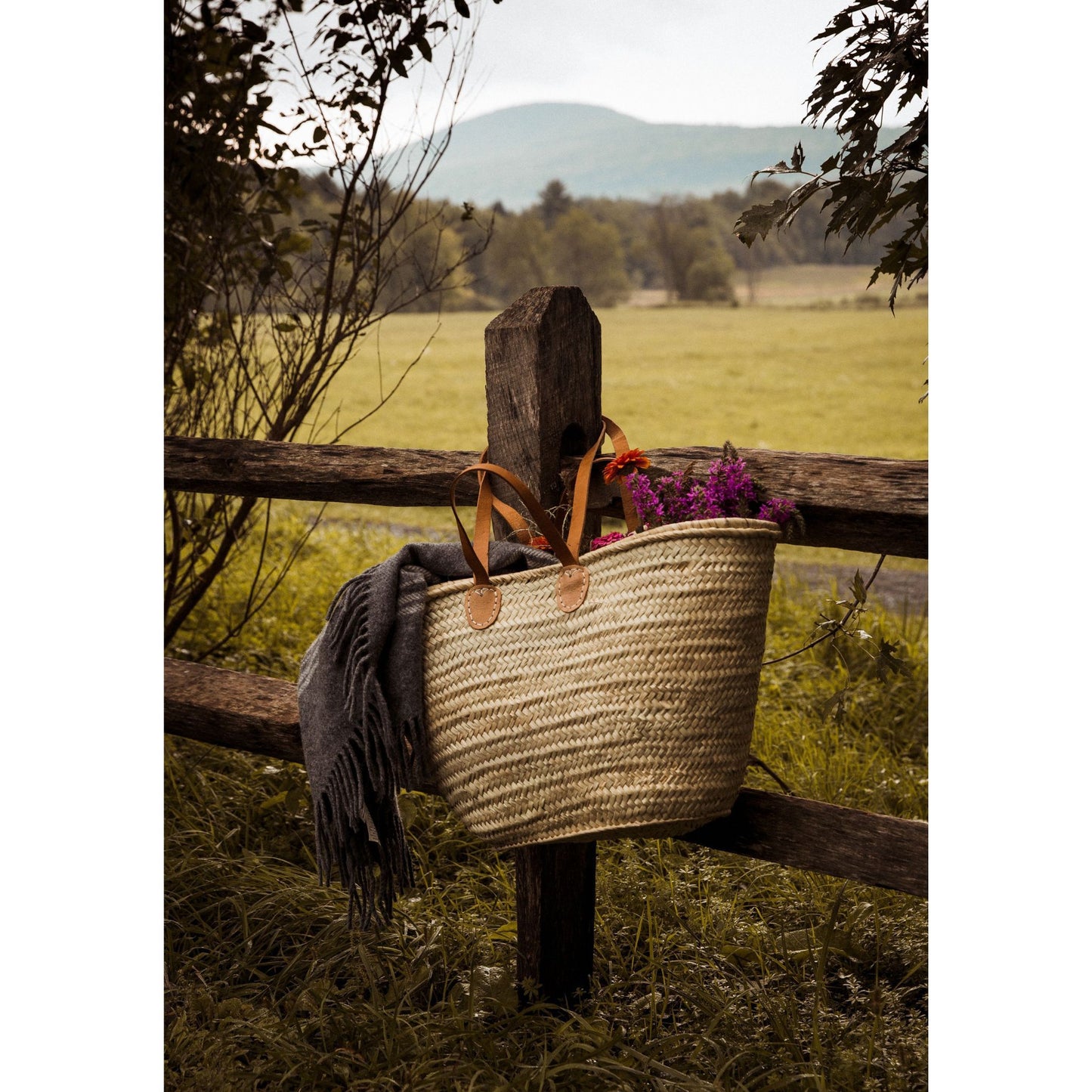 French Market Tote Basket with flowers and a blanket in it hanging on a wooden fence