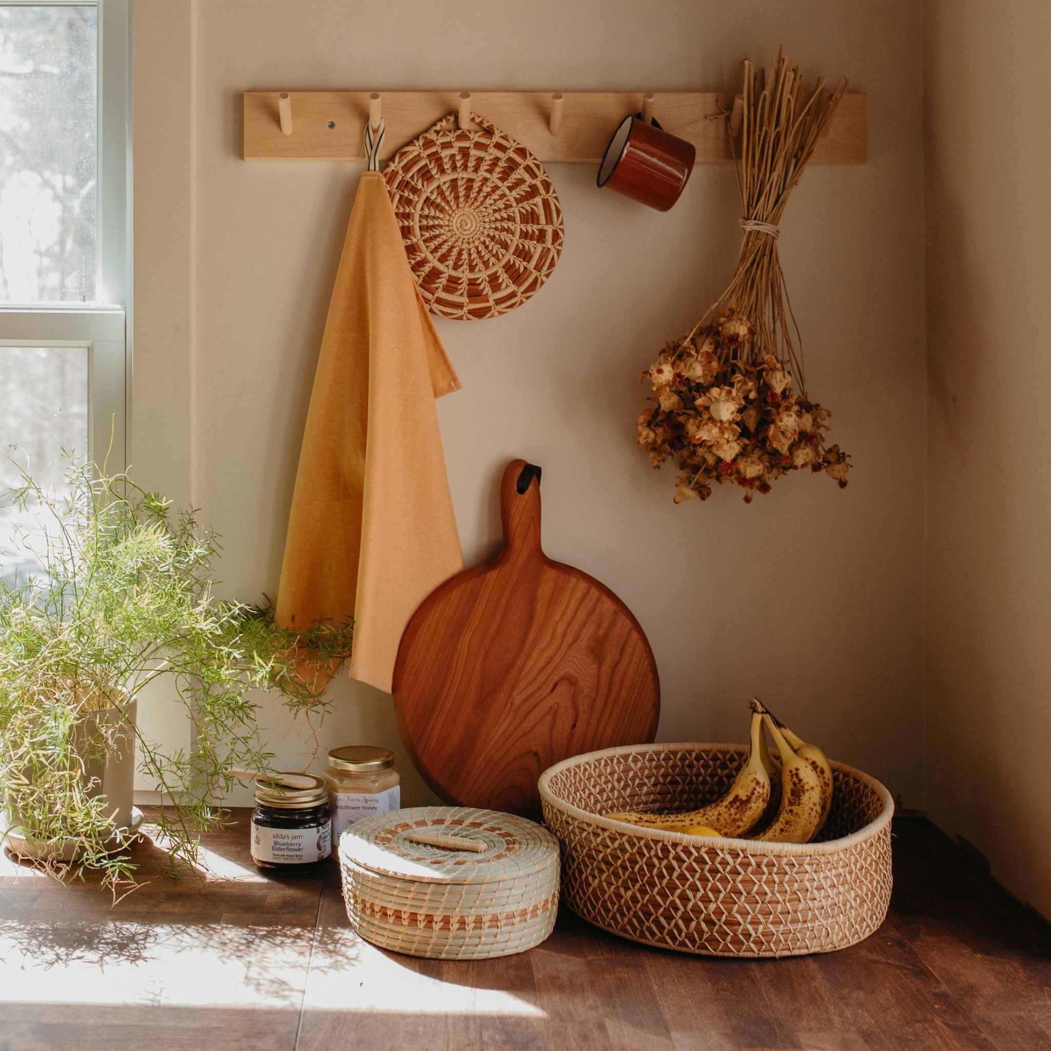 A pine needle woven trivet hanging on a wall hook next to a yellow hand towel, mug, and bouquet of dried flowers. Leaning against the wall is a cutting board and a plant, the tortilla basket is in the middle next to a larger basket filled with fruit.