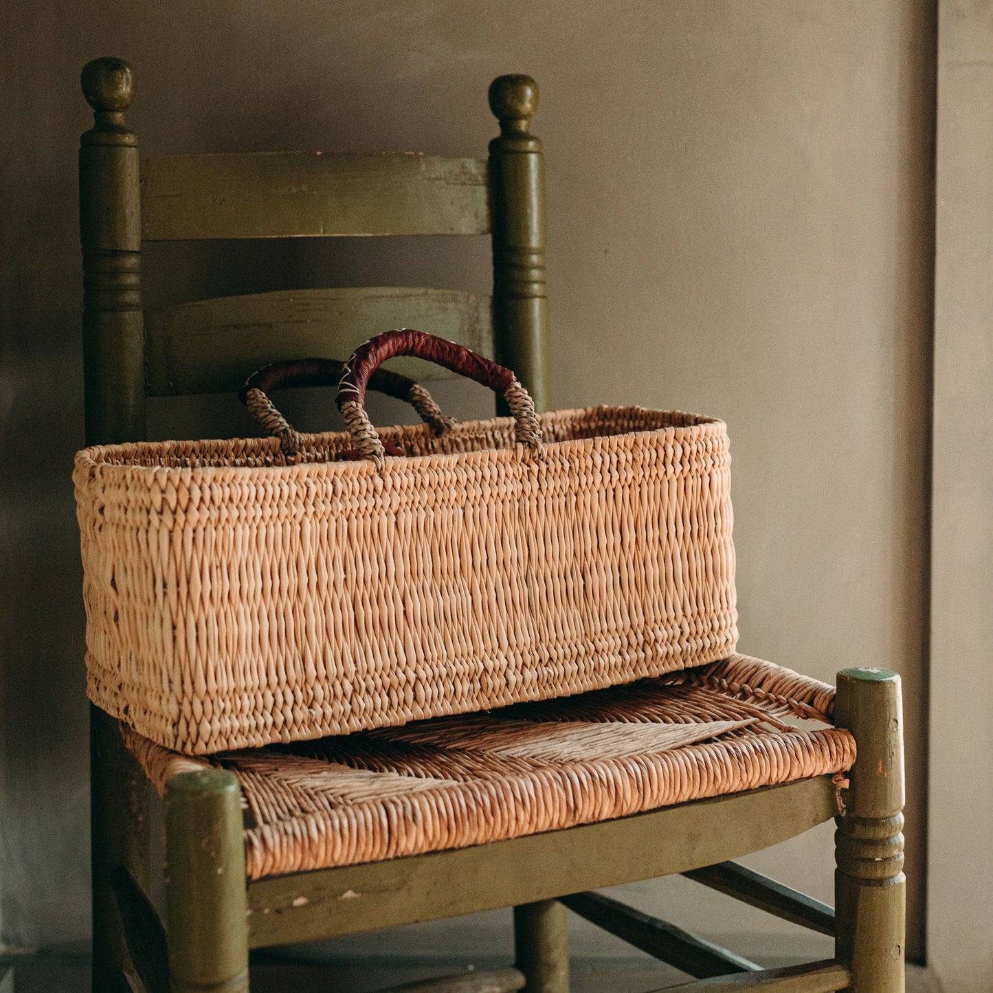 Narrow rectangular handled basket with tan leather handles sitting on a vintage chair