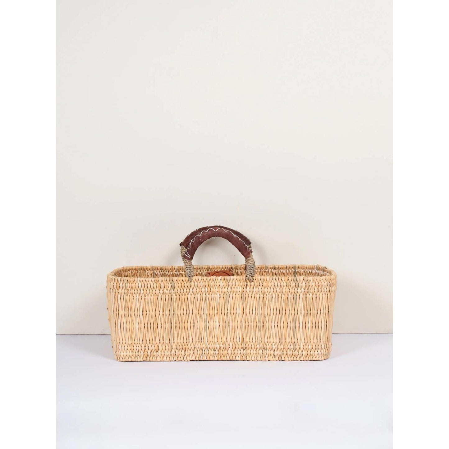 Narrow rectangular handled basket handmade in Morocco with ethically-sourced woven palm leaves and naturally tanned leather shown in medium size