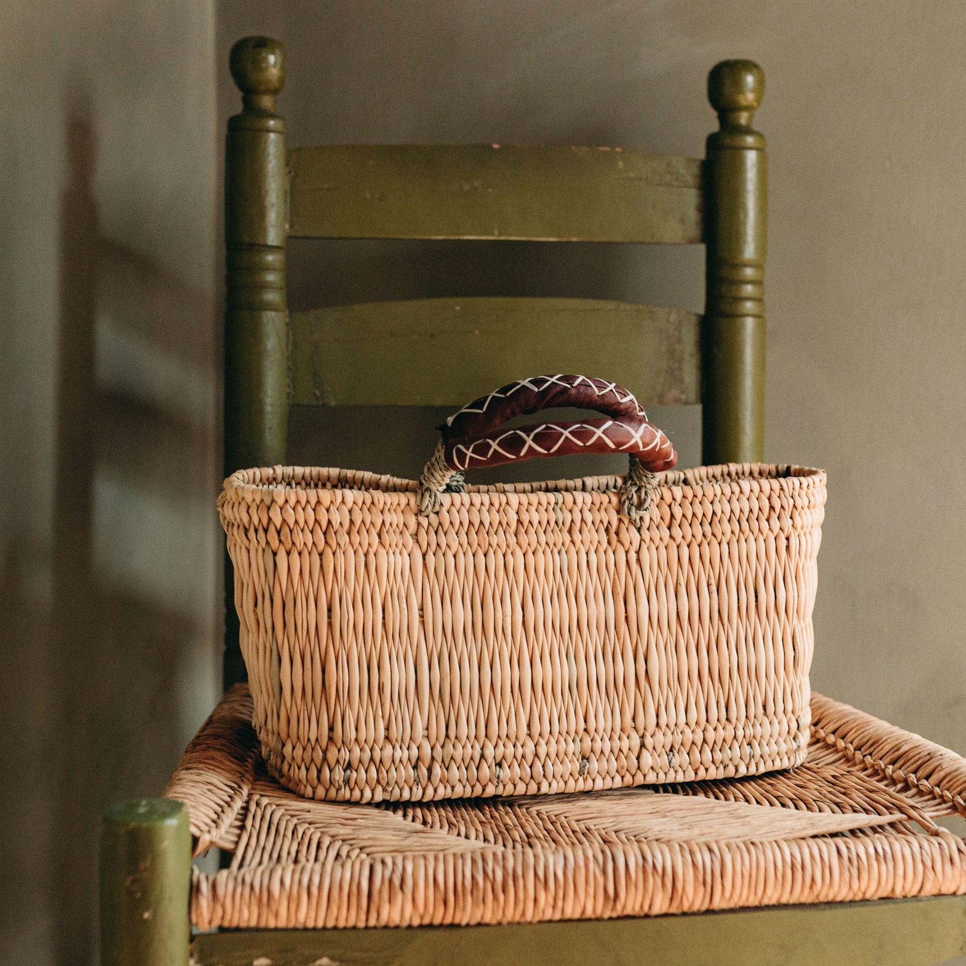 Narrow rectangular handled basket with tan leather handles sitting on a vintage chair